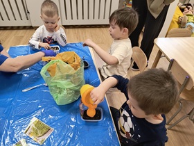3 chłopców siedzi przy stole. Każdy z nich przed sobą ma małe, plastikowe pudełko wypełnione ziemią. Na środku stolika stoi worek z ziemią. Dwóch chłopców sypie nasiona do swoich pojemników. Jeden z nich podlewa ziemię.