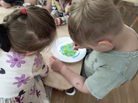 Dwoje dzieci siedzi przy stoliku. Na stole stoi talerz wypełniony sodą zabarwioną w części niebieską i zieloną farbą. Chłopiec trzyma w ręku zakraplacz, którym barwi sodę.