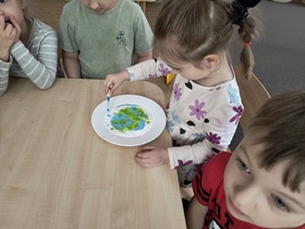 Czworo dzieci siedzi przy stoliku. Na stole stoi talerz wypełniony sodą zabarwioną w części niebieską i zieloną farbą. Dziewczynka trzyma w ręku zakraplacz, którym barwi sodę na niebiesko.