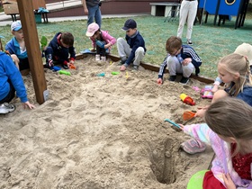 Dzieci siedzą wokół piaskownicy, dziewczynka z prawej strony zdjęcia wykopała duży dół.