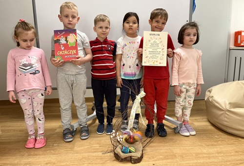 Szóstka dzieci stoi, przed nimi na podłodze ustawiony jest stroik wielkanocny. Chłopiec po lewo trzyma grę, a chłopiec po prawej stronie trzyma dyplom.