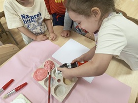 Dzieci znajdują się w sali przy stoliku. Stół wyłożony jest różowym materiałem, na którym leżą pędzle, kartka oraz pojemnik na farby. W pojemniku znajduje się 6 otworów. 2 z nich są wypełnione czerwoną mieszanką farby, kleju i pianki do golenia. Dziewczynka wyciska piankę do golenia do pustego otworu.