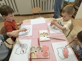 Czworo dzieci siedzi w sali przy stoliku. Stół wyłożony jest różowym materiałem, na którym leżą pędzle oraz 2 pojemniki na farby. Dzieci przed sobą mają kartki z narysowanym konturem Polski, który malują na biało i czerwono stworzonymi wcześniej farbami.