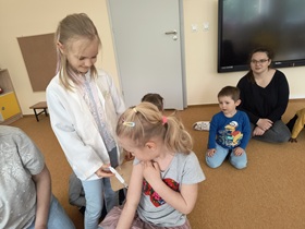 Dzieci i nauczycielki znajdują się w sali. Siedzą w kole. Przed nauczycielka znajdują się dwie dziewczynki. Jedna siedzi na krześle, druga ubrana w biały lekarski fartuch stoi obok niej, trzymając w ręku plastikową strzykawkę i udaje, że robi zastrzyk.