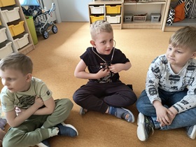 Trzech chłopców siedzi na dywanie. Chłopiec w czarnej koszulce, siedzący w środku ma założony stetoskop i słucha swojego serca.