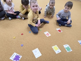 Dzieci siedzą w sali na dywanie. Przed nimi znajdują się kolorowe klocki oraz ilustracje z podpisanymi kolorami.