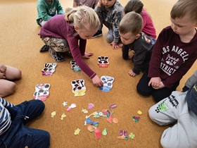Dzieci siedzą w kole na dywanie. W środku leży 6 kolorowych, papierowych słoików i wycięte kolorowe elementy. Dzieci dopasowują elementy do odpowiednich słoików.