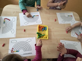 Sześcioro dzieci siedzi przy stoliku. Przed nimi znajdują się kartki z narysowaną tęczą i pudełko z plasteliną. Dzieci urywają plastelinę i wylepiają jej fragmentami tęczę.