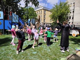 Grupa dzieci stoi na trawie na placu przedszkolnym. Dzieci mają rozłożone ręce na boki, przed nimi stoi nauczycielka.
