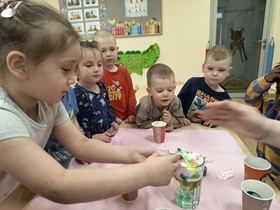 Dzieci stoją przy stoliku i obserwują, co robi koleżanka. Na środku stoi szklanka wypełniona wodą oraz pianka do golenia, zabarwiona barwnikami. Obok stoją kubki z zabarwioną wodą. Dziewczynka za pomocą strzykawki zabarwia piankę na czerwono.