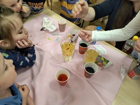 Dzieci siedzą przy stoliku, na którym stoją kubki z zabarwioną wodą oraz szklanka z wodą i olejem. Jedno z dzieci za pomocą strzykawki wpuszcza do oleju niebieski barwnik. Pozostałe dzieci obserwują powstałe zjawisko.
