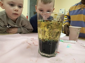 Trzech chłopców siedzi przy stoliku i przyglądają się szklance, w której znajduje się olej, zabarwiona woda oraz tabletka musująca.