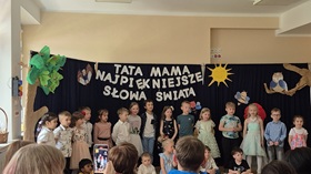Kilkanaście dzieci ustawionych w rzędzie w sali, za nimi dekoracja z napisem: Tata, mama, najpiękniejsze słowa świata.