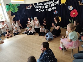 Siedmioro osób dorosłych siedzi na podłodze w półokręgu, przed każdą z tych osób siedzi dziecko, w tle za nimi dekoracja z napisem: Tata, mama, najpiękniejsze słowa świata.
