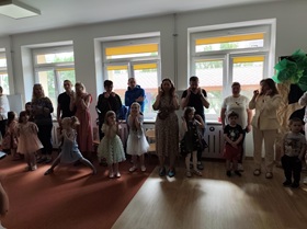 Dziewięcioro dorosłych osób stoi i wykonuje ruchy rękami, przed nimi ustawione są dzieci, za nimi w tle są trzy okna.