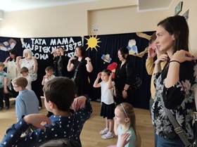 Siedmioro dorosłych osób stoi i trzyma ręce na barkach, przed każdą osobą stoi dziecko i wykonuje ten sam ruch. W tle dekoracja na granatowym tle.