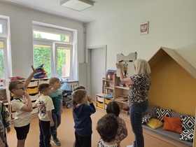 Dzieci stoją w sali. Przed nimi stoi nauczycielka, która w ręku trzyma planszę, przedstawiająca rower i razem z dziećmi omawia jego budowę.