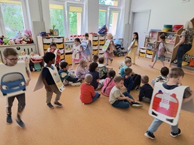 Część dzieci siedzi na dywanie. Pozostałe dzieci mają na sobie plansze, przedstawiające pojazdy i poruszają się wokół siedzących kolegów i koleżanek.