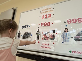 Na tablicy przywieszony jest napis "Numery alarmowe". Poniżej są numery: 997, 998, 999.
Pod numerami przywieszone są ilustracje, przedstawiające policjanta, wóz policyjny, strażaka, wóz strażacki, lekarkę, ambulans. Pod nimi znajdują się podpisy. Przed tablicą stoi dziewczynka, która palcem wskazuje na strażaka.