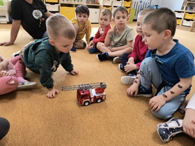 Dzieci siedzą w kole na dywanie. W środku stoi wóz strażacki.