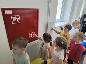 Dzieci stoją na korytarzu przed skrzynką, zawierająca gaśnicę i wąż strażacki.