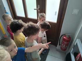 Dzieci stoją na korytarzu przy drzwiach wejściowych do przedszkola. Oglądają gaśnice i znajdujące się w jej pobliżu oznaczenia.