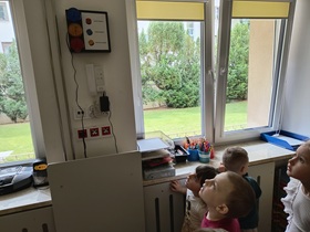 Dzieci stoją w sali i przypatrują się sygnalizatorowi, umieszczonemu na ścianie. Niebieski - alarm napadowy, żółty - odwołanie alarmu, czerwony - alarm pożarowy.