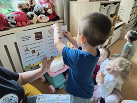 Dzieci siedzą na podłodze w sali. Przed nimi siedzi nauczycielka, która trzyma w ręku książkę o misiu, odwiedzającym remizę strażacką. Chłopiec stoi obok i przykleja naklejkę w wyznaczone miejsce.