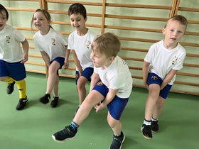 Piątka dzieci w strojach sportowych wykonuje ćwiczenia – stojąc podnoszą jedną nogę.
