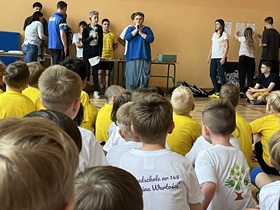 Dzieci w kolorowych koszulkach siedzą na podłodze. Przed nimi stoi osoba dorosła z mikrofonem.