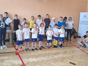 Szóstka dzieci w białych koszulkach i niebieskich spodenkach stoi w rzędzie, na szyi mają zawieszone medale. Pośrodku stoi dziewczynka z uniesionymi rękoma, w których trzyma puchar. Za nimi stoi 11 dorosłych osób.