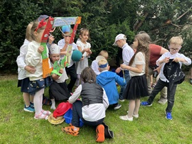 Chłopcy i dziewczynki zgromadzone są wokół koszyka, z którego wyjmują zgromadzone rzeczy: czapki, okulary, maski. Dziewczynki stoją i trzymają ramkę do foto budki.