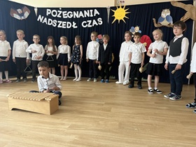 Na tle dekoracji ustawione są dzieci, w dłoniach trzymają instrumenty: trójkąty, bębenki grzechotki, kołatki. Przed dziećmi na podłodze siedzi chłopiec, przed nim ustawione są cymbałki.