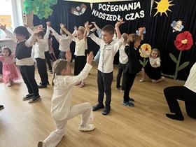 Dzieci ustawione są parami w kole. Dziewczynki klęczą na jednym kolanie, ręce mają uniesione w górę. Chłopcy stoją przed nimi, ręce obracają przed sobą.