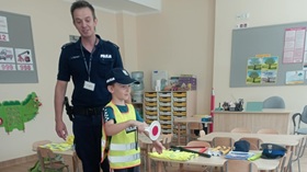 Policjant stoi, przed nim chłopiec, przebrany za funkcjonariusza policji. Chłopiec ubrany jest w odblaskową kamizelkę z napisem policja, na głowie ma policyjną czapkę, a w ręku trzyma policyjnego lizaka.
