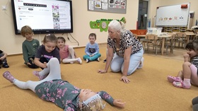 Dzieci siedzą w kole. W środku koła na dywanie leży dziewczynka i udaje poszkodowaną. Druga dziewczynka klęczy przy jej nogach i próbuje ułożyć ją w pozycji bocznej bezpiecznej. Nauczycielka znajduje się obok i próbuje wesprzeć dziewczynkę.