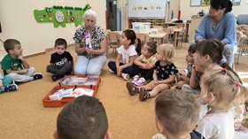 Dzieci i panie siedzą w kole na dywanie. W środku koła znajduje się otwarta, rozłożona apteczka przedszkolna. Nauczycielka prezentuje dzieciom zawartość apteczki.