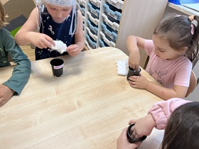 Przy stoliku siedzi czworo dzieci. Dwie dziewczynki wkładają dłonie do małych, czarnych pojemników. Dziewczynka naprzeciwko trzyma w dłoniach watę, przed nią stoi mała doniczka.