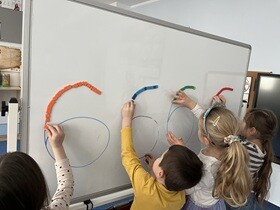 Przed białą tablicą stoi czworo dzieci, przed każdym napisana jest cyfra 6, którą ozdabiają małymi kolorowymi klockami magnetycznymi.