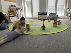 Na dywanie leży dziewczynka, z jej lewej strony ukucnął chłopiec. Przed nimi ustawione są drewniane sześciany z czarnymi sześcioma otworami.