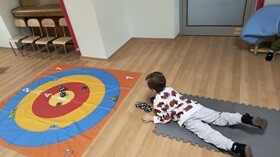 Chłopiec leży na brzuchu na piankowych puzzlach i rzuca materiałowymi piłeczkami do rozłożonej, kolorowej maty.