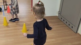 2 dziewczynki na sali gimnastycznej. Dzieci pod stopami mają kolorowe woreczki i jadą na nich jak na nartach omijając pachołki.