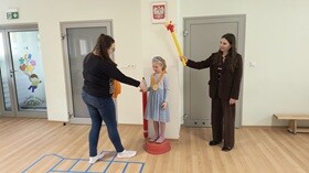 Dziewczynka stoi na podeście. Na szyi powieszony na medal wykonany z papieru i bibuły. Obok dziewczynki stoją 2 nauczycielki. Jedna z prowadzących gratuluję dziewczynce a druga trzyma uniesiony papierowy znicz olimpijski.