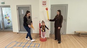 Dziewczynka stoi na podeście, uśmiecha się i ręce ma rozłożone na boki. Na szyi ma powieszony medal wykonany z papieru i bibuły. Obok dziewczynki stoją 2 nauczycielki. Jedna z prowadzących gratuluję dziewczynce a druga trzyma uniesiony papierowy znicz olimpijski.