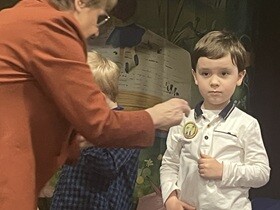 Pani w brązowej marynarce zawiesza przypinkę na bluzce chłopca.