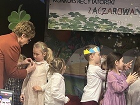 4 Pani w brązowej marynarce przypina dziewczynce przypinkę. Z prawej strony dzieci wymachują rękoma w stronę baniek.