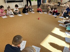 Dzieci siedzą w kole, przed nimi leżą kartki z narysowanymi rysunkami.