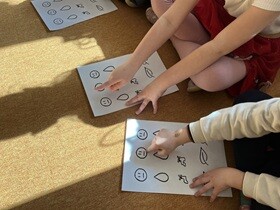 Na dywanie siedzi dwoje dzieci, przed nimi leży kartka z rysunkami buzi, kranu i kropli. Dzieci paluszkiem wskazują narysowaną buźkę.