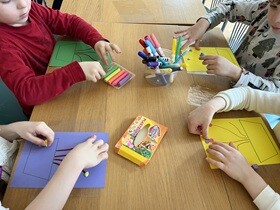 Czworo dzieci siedzi przy stole, przed nimi leżą kolorowe kartki z narysowaną donicą i łodygami. Dzieci wyklejają łodygi kolorową plasteliną.