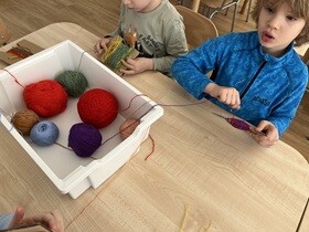 Na stole stoi biały pojemnik, w którym ułożone są kolorowe włóczki. Dwaj chłopcy nawijają wełnę na tekturkę.